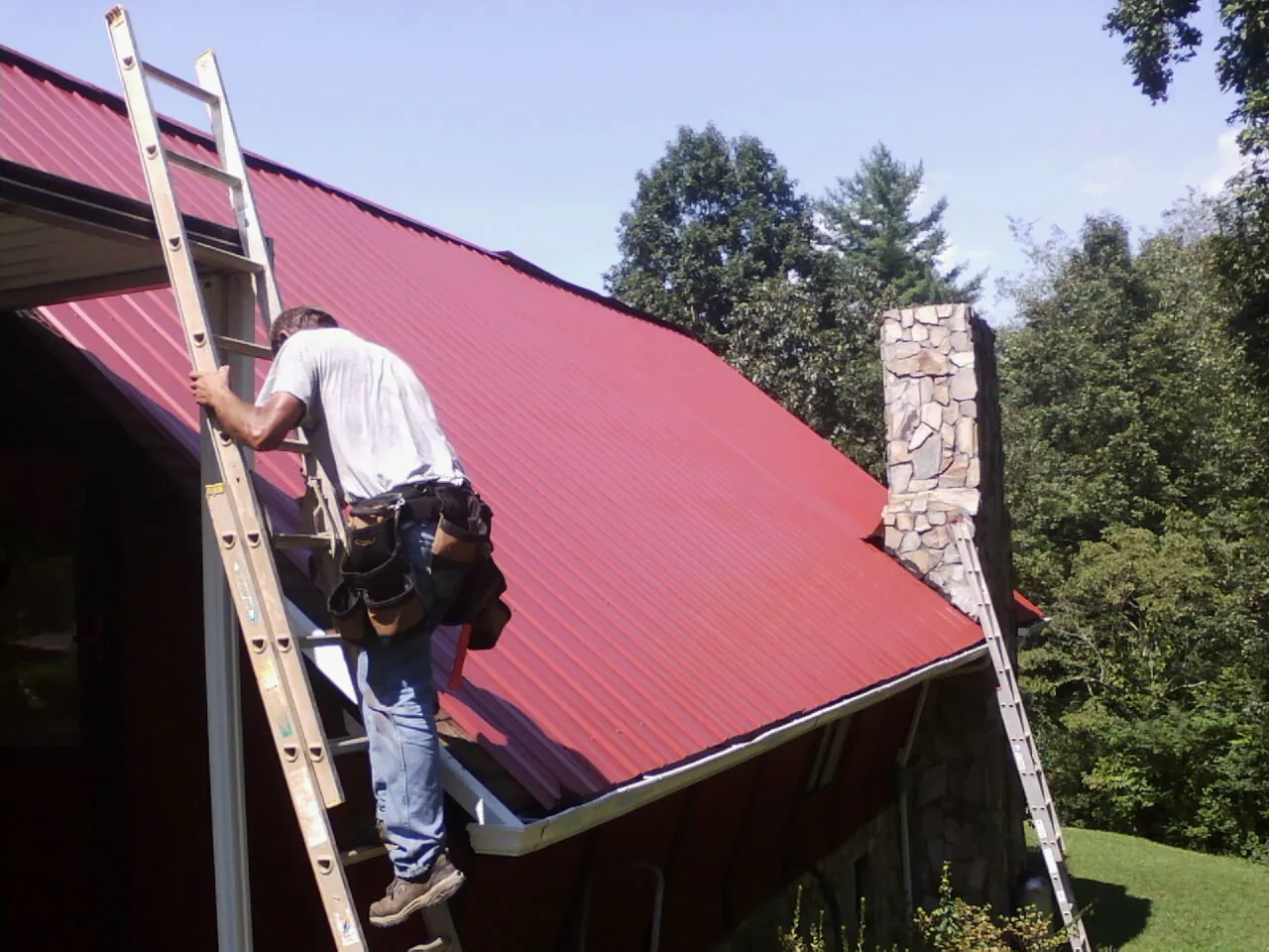 Roofer on a ladder beside a red metal roof and stone chimney — Soffit & Fascia Repair by Roofing Brothers in New Enterprise